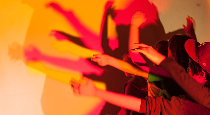 Mehrere Personen heben ihre Arme vor einer Lichtquelle und werfen überlappende, bunte Schatten auf eine Wand.