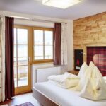 Ein modernes Hotelzimmer mit einem Doppelbett, einem holzgetäfelten Kopfteil, kariertem Bettzeug, einem an der Wand montierten Fernseher und einem Fenster mit Vorhängen, die auf einen Balkon führen.