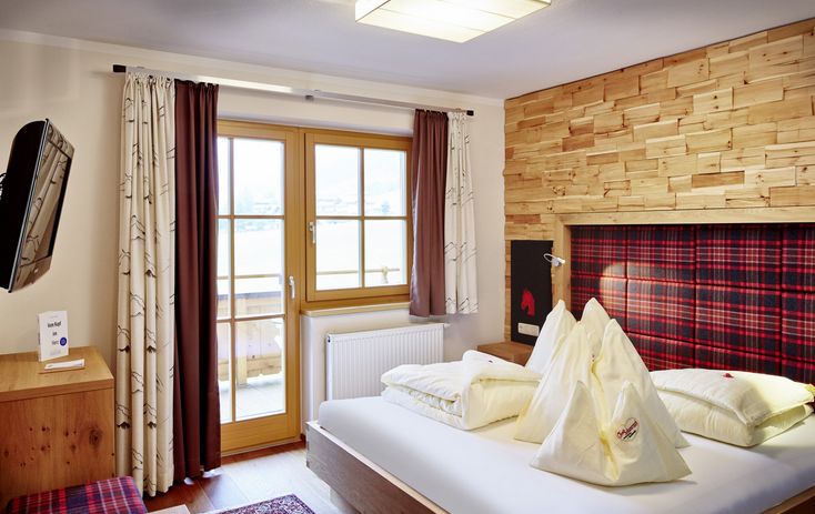 Ein modernes Hotelzimmer mit einem Doppelbett, einem holzgetäfelten Kopfteil, kariertem Bettzeug, einem an der Wand montierten Fernseher und einem Fenster mit Vorhängen, die auf einen Balkon führen.