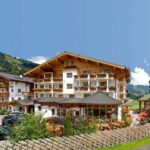 Ein großes Alpenhotel mit Holzbalkonen und Blumenkästen, umgeben von Bergen und Grün unter einem klaren blauen Himmel. Mehrere Autos sind draußen geparkt.