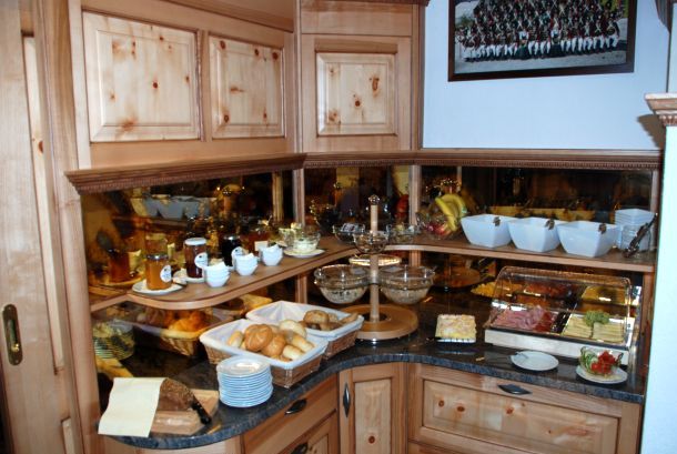 Ein Frühstücksbuffet mit Brötchen, Aufschnitt, Käse, Obst, Joghurt, Bechern und Gläsern, die auf Holzregalen und -theken angeordnet sind.