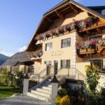 Ein zweistöckiges Gebäude im Chalet-Stil mit Balkonen, die mit bunten Blumenkästen geschmückt sind, einer Eingangstreppe und einem Garten mit Sonnenblumen im Vordergrund.