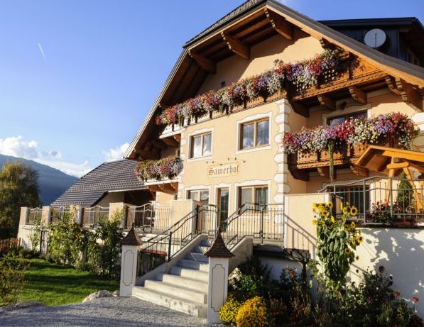 Ein zweistöckiges Gebäude im Chalet-Stil mit Balkonen, die mit bunten Blumenkästen geschmückt sind, einer Eingangstreppe und einem Garten mit Sonnenblumen im Vordergrund.