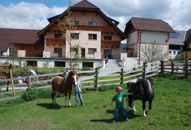 Zwei Kinder stehen auf einer eingezäunten Wiese mit zwei kleinen Pferden vor einem Bauernhaus und anderen Gebäuden unter einem teilweise bewölkten Himmel.