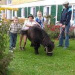 Vier Kinder stehen in der Nähe eines kleinen schwarzen Ponys, das im Gras grast, während ein Erwachsener seine Leine hält. Im Hintergrund ist ein Gebäude mit grünen Fensterläden und einem Garten zu sehen.