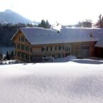 Ein großes Holzgebäude, aus dessen Schornstein Rauch aufsteigt, ist umgeben von tiefem Schnee und Bäumen in einer bergigen Winterlandschaft.