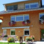 Ein dreistöckiges Haus mit Backsteinfassade und Obergeschoss aus Holz, mit Blumenkästen unter den Fenstern und Topfpflanzen auf der Terrasse.