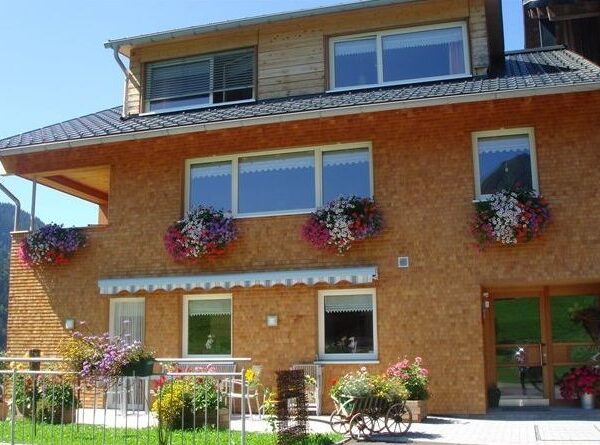 Ein dreistöckiges Haus mit Backsteinfassade und Obergeschoss aus Holz, mit Blumenkästen unter den Fenstern und Topfpflanzen auf der Terrasse.
