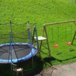 Ein Trampolin mit einem Sicherheitsnetz, zwei Schaukeln, eine Strickleiter und eine Bank sind auf einer Wiese neben einem abschüssigen Hügel aufgestellt.