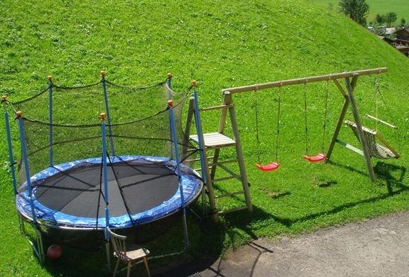 Ein Trampolin mit einem Sicherheitsnetz, zwei Schaukeln, eine Strickleiter und eine Bank sind auf einer Wiese neben einem abschüssigen Hügel aufgestellt.