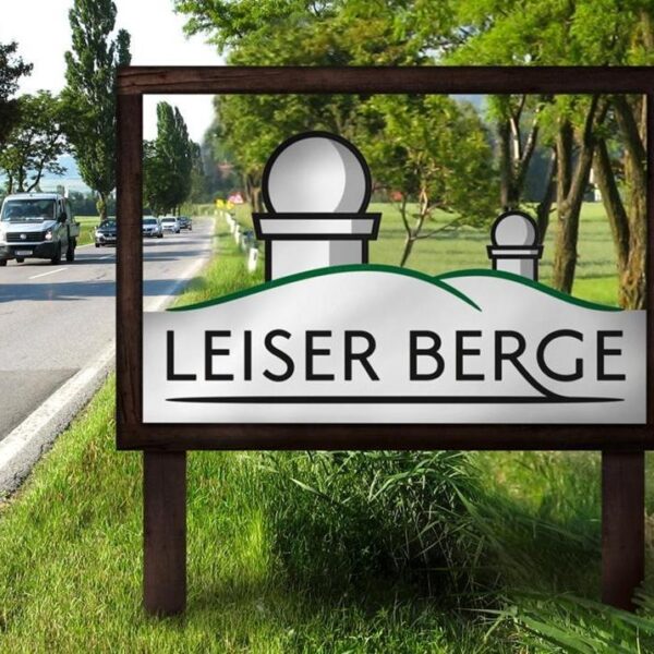 Auf einem Straßenschild steht „Leiser Berge“ und die Grafik zeigt zwei Hügel und zwei Observatoriumskuppeln. Im Hintergrund sind Bäume und ein Lieferwagen zu sehen.
