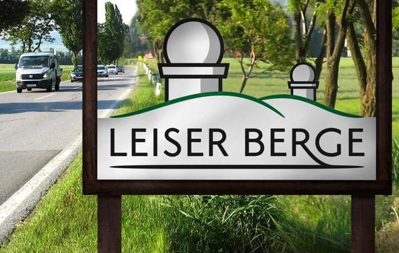 Auf einem Straßenschild steht „Leiser Berge“ und die Grafik zeigt zwei Hügel und zwei Observatoriumskuppeln. Im Hintergrund sind Bäume und ein Lieferwagen zu sehen.