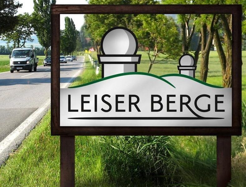 Auf einem Straßenschild steht „Leiser Berge“ und die Grafik zeigt zwei Hügel und zwei Observatoriumskuppeln. Im Hintergrund sind Bäume und ein Lieferwagen zu sehen.