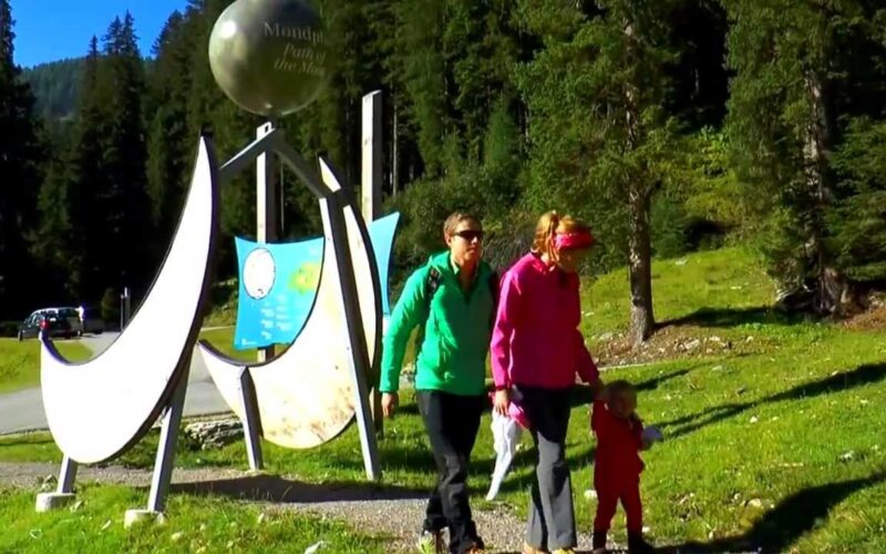 Drei Personen, zwei Erwachsene und ein Kind, gehen bei Tageslicht auf einem Graspfad in der Nähe einer großen Außenskulptur und eines Informationsschildes, umgeben von Bäumen.