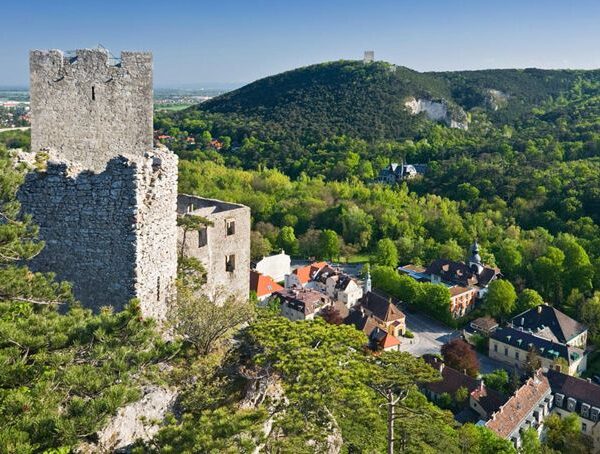 Die Steinruinen einer Burg auf einem Hügel überblicken ein kleines Dorf mit bunten Häusern, umgeben von dichten grünen Wäldern und fernen Hügeln unter einem klaren Himmel.