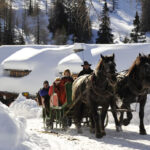Ein Pferdeschlitten mit mehreren Personen fährt auf einem verschneiten Weg mit schneebedeckten Bäumen und einer Hütte im Hintergrund.