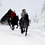 Zwei Personen reiten auf einem verschneiten Pfad neben einer Holzhütte, gefolgt von einem Hund, mit schneebedeckten Bäumen und einem Zaun im Hintergrund.