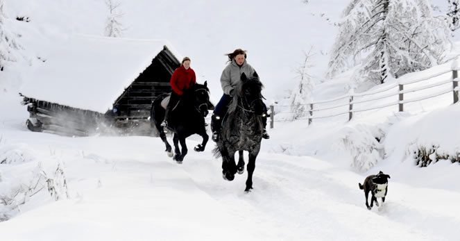 Zwei Personen reiten auf einem verschneiten Pfad neben einer Holzhütte, gefolgt von einem Hund, mit schneebedeckten Bäumen und einem Zaun im Hintergrund.
