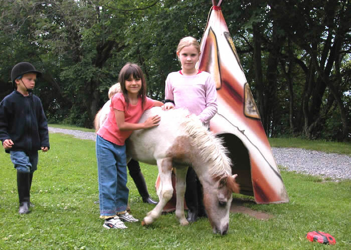 Drei Kinder stehen neben einem kleinen Pony, das vor einem bemalten Tipi-Gebäude auf der Wiese grast. Im Hintergrund sind Bäume und ein Schotterweg zu sehen.
