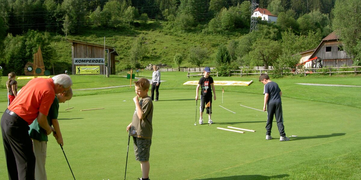 Eine Gruppe von Menschen, darunter Kinder und Erwachsene, übt das Putten auf einem von Bäumen und Gebäuden umgebenen Golfgrün in einer ländlichen Gegend.