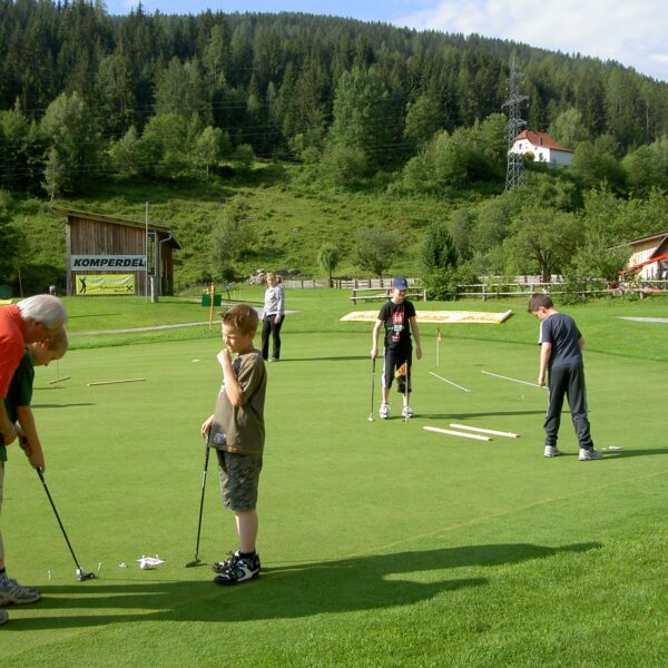 Eine Gruppe von Menschen, darunter Kinder und Erwachsene, übt das Putten auf einem von Bäumen und Gebäuden umgebenen Golfgrün in einer ländlichen Gegend.