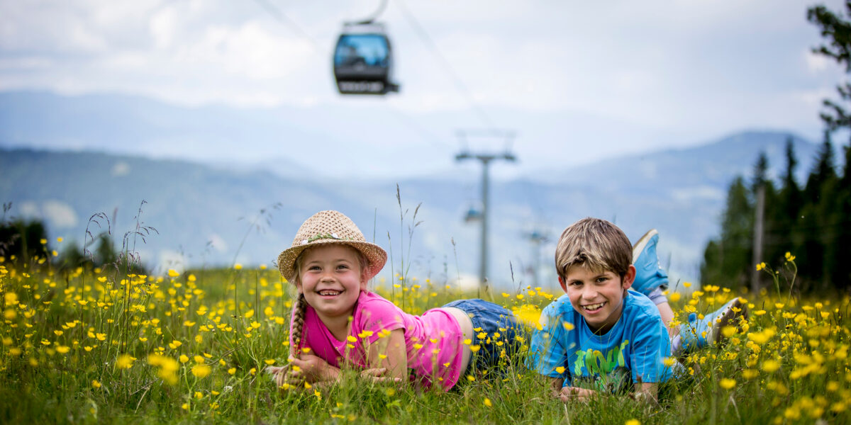 Zwei Kinder liegen auf dem Bauch in einem Feld mit gelben Wildblumen und lächeln in die Kamera, im Hintergrund sind Berge und eine Seilbahn zu sehen.
