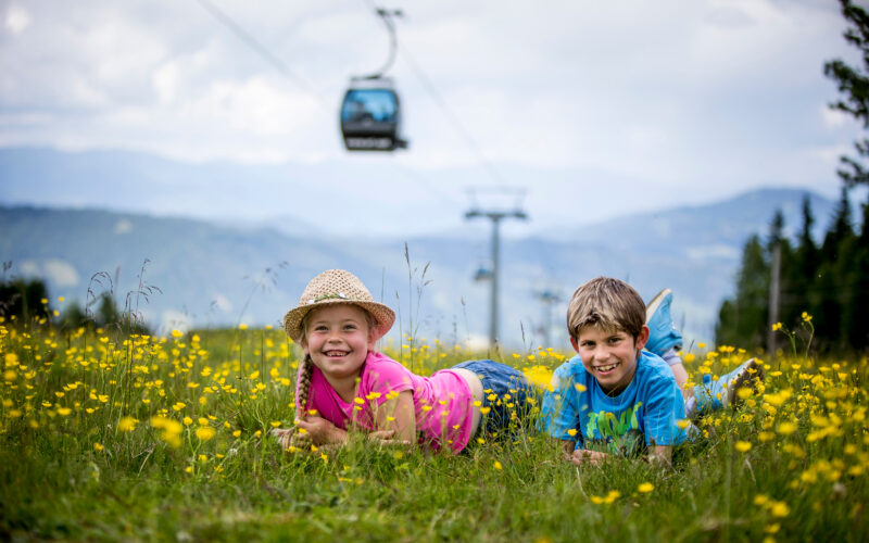 Zwei Kinder liegen auf dem Bauch in einem Feld mit gelben Wildblumen und lächeln in die Kamera, im Hintergrund sind Berge und eine Seilbahn zu sehen.