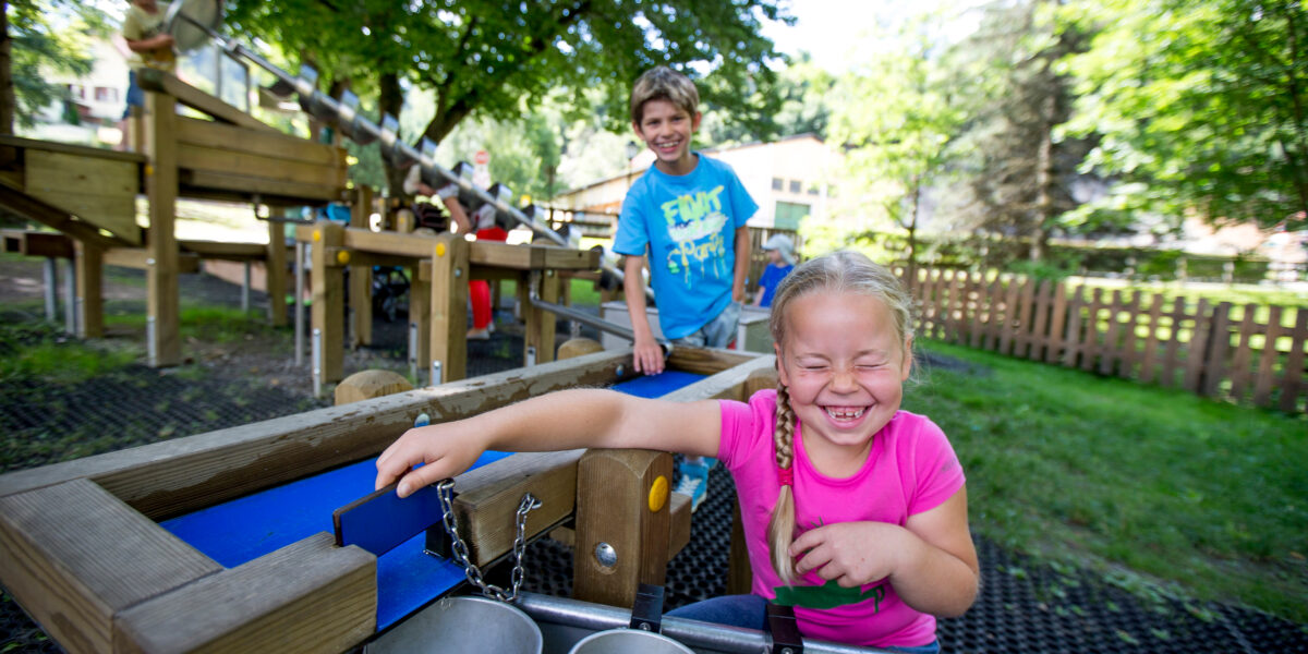 Zwei Kinder spielen an einer Wasseraktivitätsstation auf einem Spielplatz im Freien, im Hintergrund sind Bäume und Holzgeräte zu sehen.