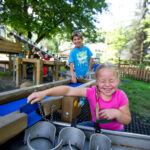 Zwei Kinder spielen an einer Wasseraktivitätsstation auf einem Spielplatz im Freien, im Hintergrund sind Bäume und Holzgeräte zu sehen.