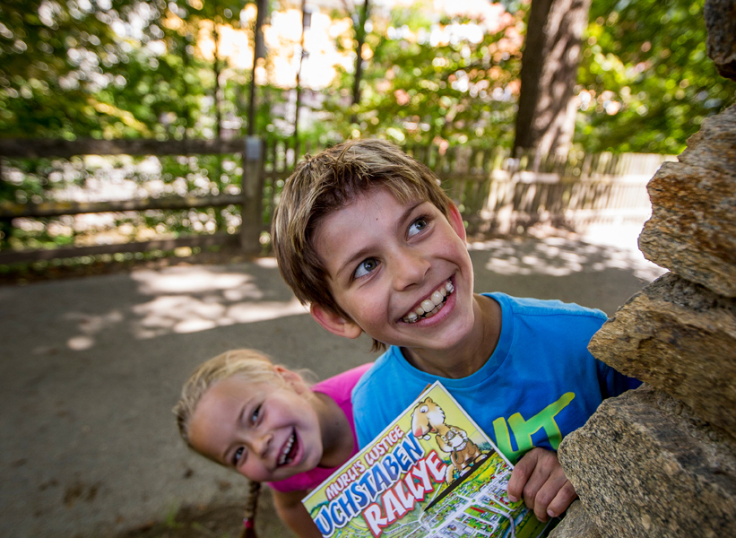 Zwei lächelnde Kinder stehen im Freien und spähen um eine Steinmauer herum. Der Junge hält eine bunte Karte mit der Aufschrift "Rätsel-Rallye" in der Hand. Im Hintergrund sind Bäume und ein Zaun zu sehen.