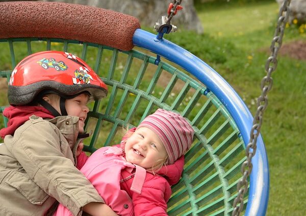 Zwei kleine Kinder in warmer Kleidung liegen zusammen auf einer runden, blauen Schaukel im Freien. Eines trägt einen Helm, beide lächeln. Im Hintergrund sind Gras und Spielgeräte zu sehen.