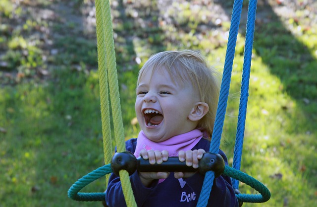 Ein kleines Kind mit blondem Haar lacht, während es sich im Freien an den Seilen einer Schaukel festhält.