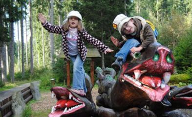Zwei Kinder mit Hüten posieren spielerisch auf einer großen Drachenskulptur in einer bewaldeten Umgebung im Freien.