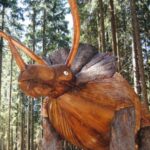 Eine große Holzskulptur eines Triceratops-Dinosauriers steht zwischen hohen Bäumen in einem Waldgebiet.