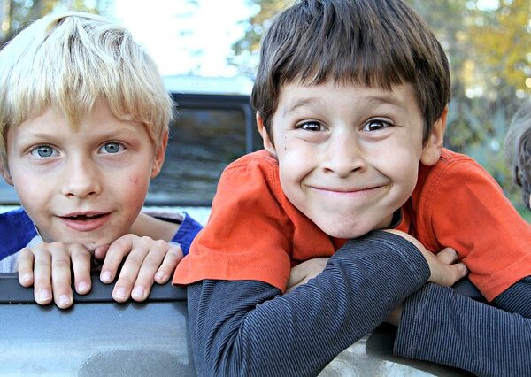 Zwei kleine Jungen lehnen sich im Freien über eine Fläche, einer mit blondem Haar und blauen Augen, der andere mit braunem Haar und einem breiten Lächeln, beide blicken in die Kamera.