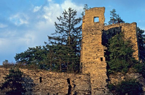 Ruinen eines alten Steinturms und einer Mauer, teilweise von Bäumen bedeckt, unter einem blauen Himmel mit vereinzelten Wolken.