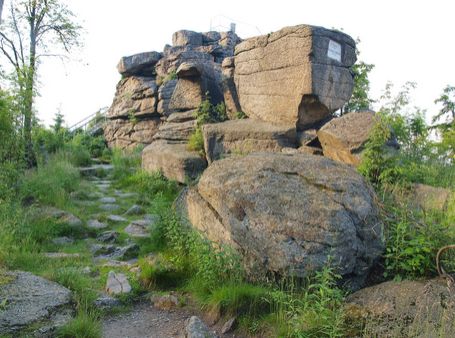 Große, von Grün umgebene Felsformationen und ein Feldweg, der zu ihnen hinaufführt, mit einem kleinen Metallgeländer, das oben sichtbar ist.