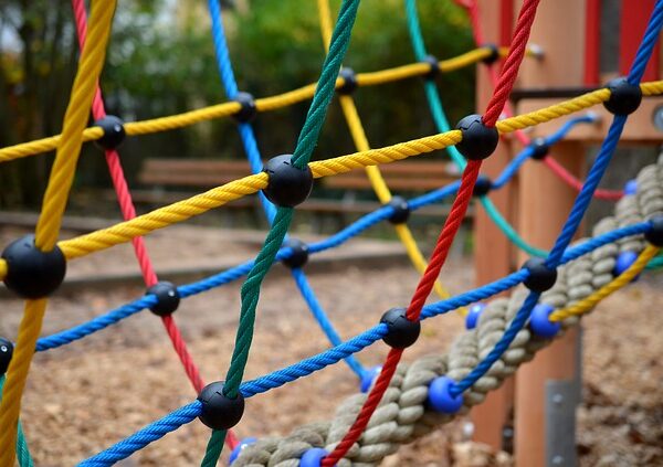 Nahaufnahme einer farbenfrohen Kletterseilnetzstruktur auf einem Spielplatz im Freien, mit Holzspänen auf dem Boden und unscharfen Bänken im Hintergrund.