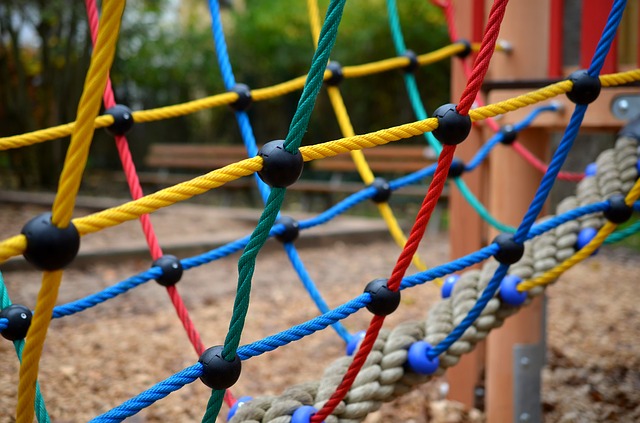 Nahaufnahme einer farbenfrohen Kletterseilnetzstruktur auf einem Spielplatz im Freien, mit Holzspänen auf dem Boden und unscharfen Bänken im Hintergrund.