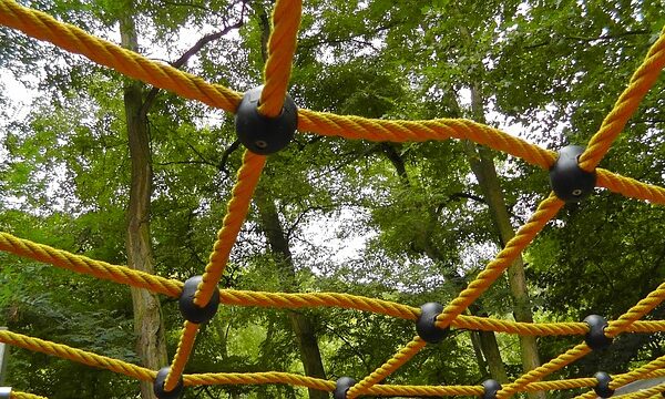 Gelbes Seilkletternetz mit schwarzen Verbindern vor einem Hintergrund aus grünen Bäumen in einem Außenbereich.