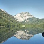 Ein klarer See spiegelt einen bewaldeten Berg unter einem blauen Himmel wider, mit felsigen Hängen und Bäumen entlang der Uferlinie.
