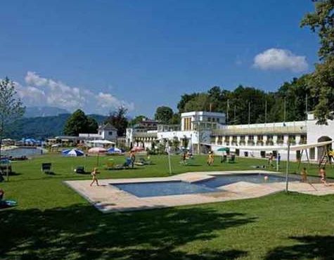 Ein grasbewachsener Erholungsbereich mit einem flachen Außenpool, entspannenden Menschen und einem weißen modernen Gebäude im Hintergrund unter einem teilweise bewölkten Himmel.