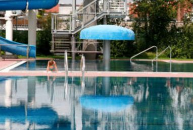 Ein Außenpool mit klarem Wasser, einem blauen, pilzförmigen Brunnen, Metallgeländern und einer Wasserrutsche. Eine Person ruht sich am Beckenrand aus.