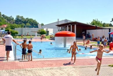 An einem sonnigen Tag erfreuen sich Kinder und Erwachsene an einem Außenpool mit einem roten, pilzförmigen Wasserspiel.