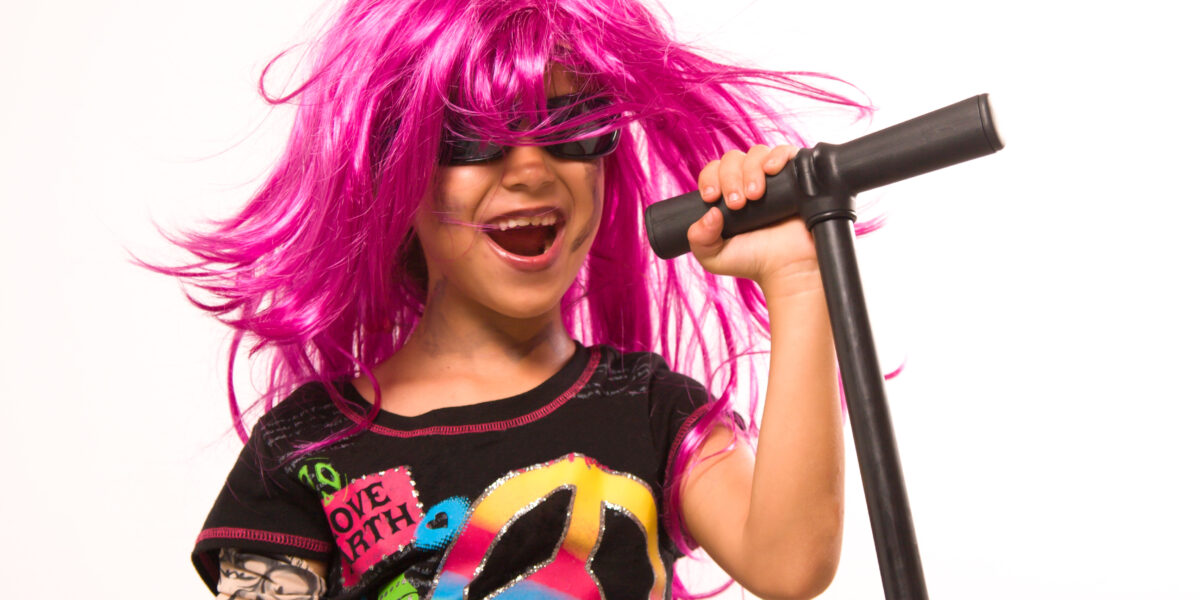 Ein Kind mit rosa Perücke und Sonnenbrille singt in ein Mikrofon, während es ein buntes T-Shirt mit Friedens- und Herzsymbolen trägt.