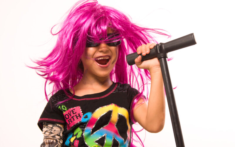 Ein Kind mit rosa Perücke und Sonnenbrille singt in ein Mikrofon, während es ein buntes T-Shirt mit Friedens- und Herzsymbolen trägt.