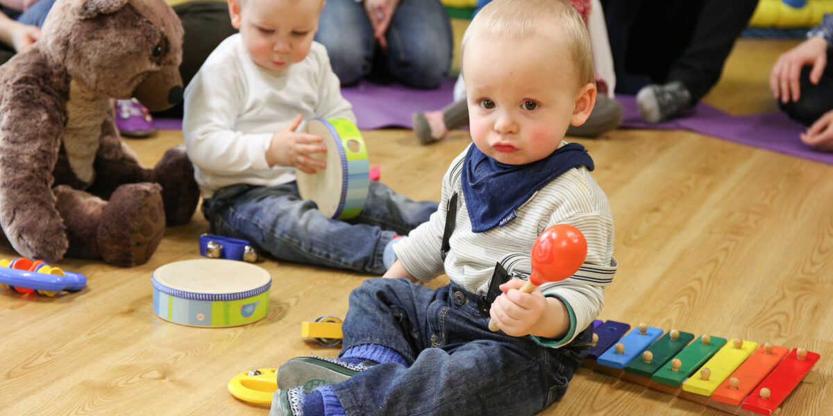 Zwei Kleinkinder sitzen auf dem Boden und spielen mit Musikinstrumenten; eines hält eine Maraca und sitzt neben einem Xylophon, während das andere eine Trommel spielt. Ein großer ausgestopfter Bär ist in der Nähe.