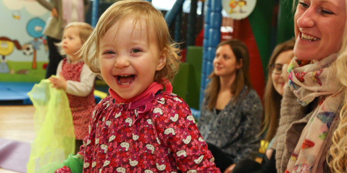 Ein kleines Kind in einem roten Kleid lächelt und lacht, umgeben von Erwachsenen und anderen Kindern in einem Indoor-Spielbereich.