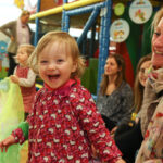 Ein kleines Kind in einem roten Kleid lächelt und lacht, umgeben von Erwachsenen und anderen Kindern in einem Indoor-Spielbereich.