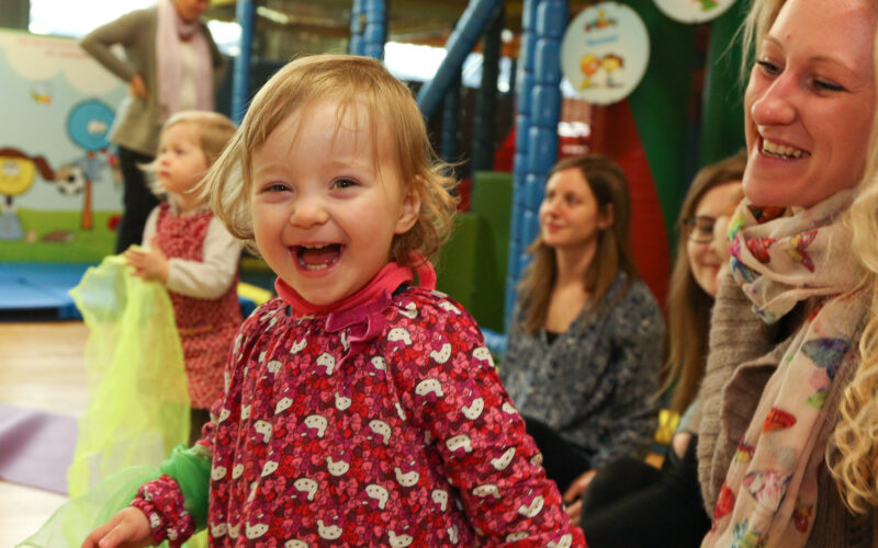 Ein kleines Kind in einem roten Kleid lächelt und lacht, umgeben von Erwachsenen und anderen Kindern in einem Indoor-Spielbereich.